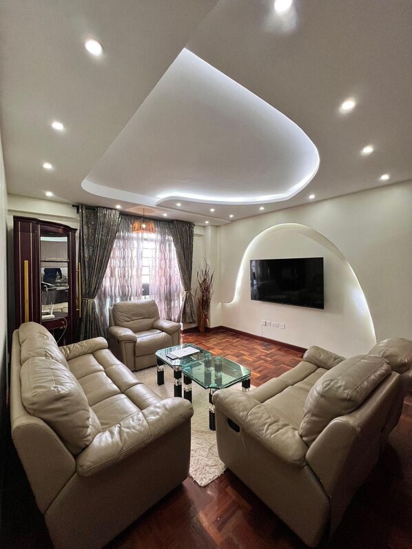 A modern living room with leather sofas, a glass coffee table, and a flat-screen TV mounted on a curved wall. - Cosy Nairobi 1bdr Apartment for Two in Westlands (Nairobi)