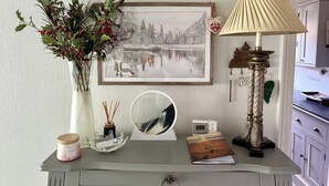A desk with a framed picture, a vase with greenery, a candle, a mirror, a lamp, and a small clock.