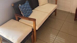 A sofa with a cushion and a matching ottoman, a woven backrest, and a window with blinds.