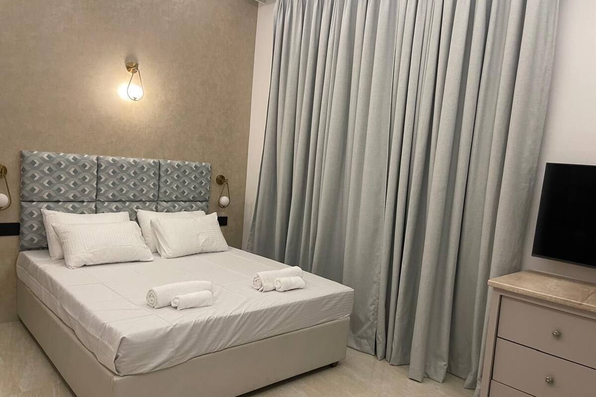 Une chambre moderne avec un grand lit, une tête de lit ornée d’un motif géométrique, une lampe de chevet et un téléviseur posé sur une commode.