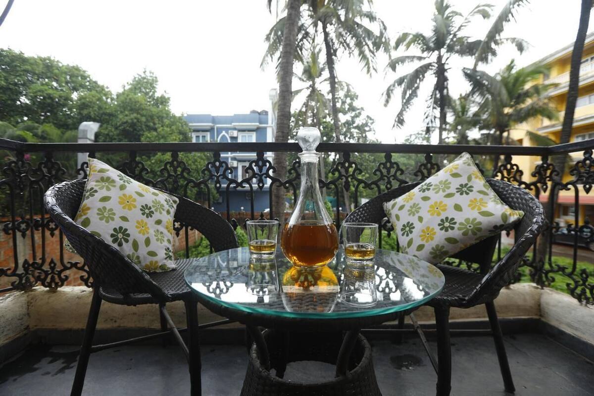 Une table en verre, deux chaises et une bouteille de spiritueux, disposés sur un balcon doté d’une rambarde en métal et avec des palmiers en arrière-plan.