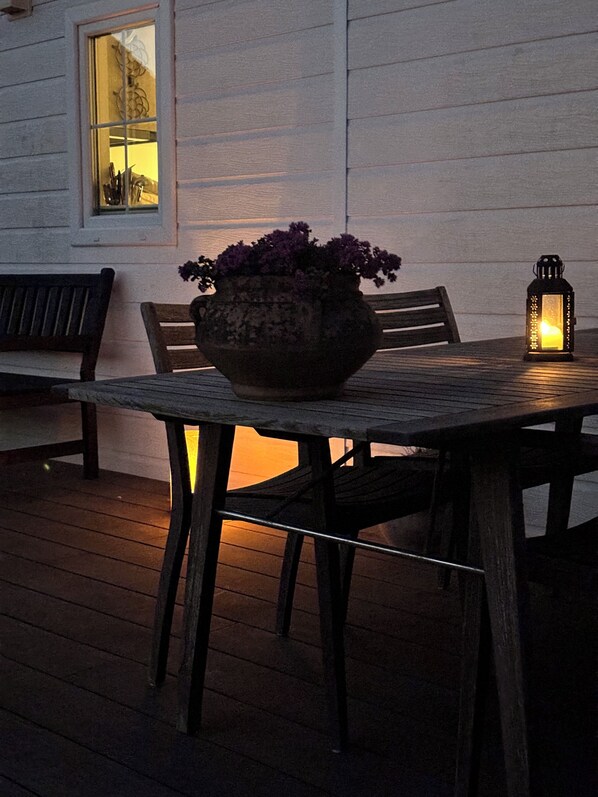 Una terraza de madera con una mesa, sillas, una planta en maceta y un farol.