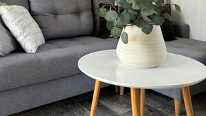 Un sofá gris con un cojín blanco, una mesa blanca con patas de madera y una planta en maceta con hojas verdes.