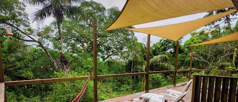 A wooden deck with white lounge chairs and a canopy, overlooking a lush garden with palm trees.
