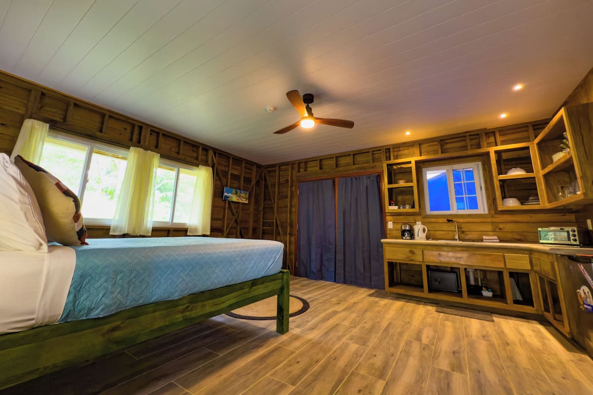 Una habitación de cabaña de madera con cama, ventilador de techo, ventana con cortinas, una pequeña cocina y televisión.