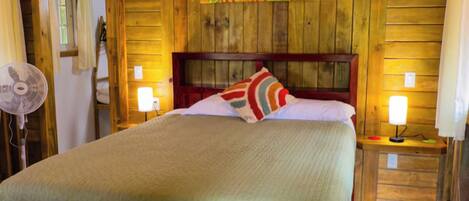 A wooden-framed room with a bed, a fan, and a wall painting.