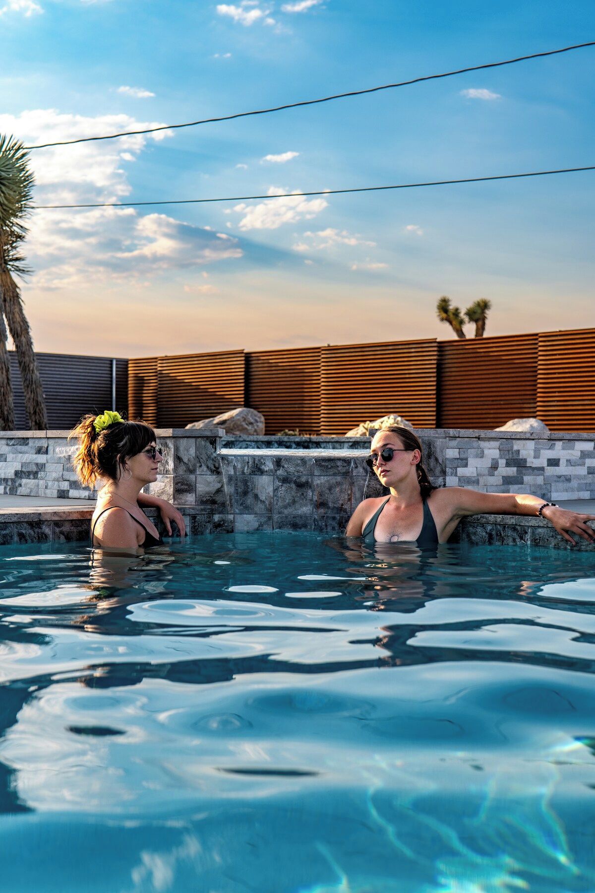 A swimming pool with two people in it, surrounded by a stone wall and a wooden fence.