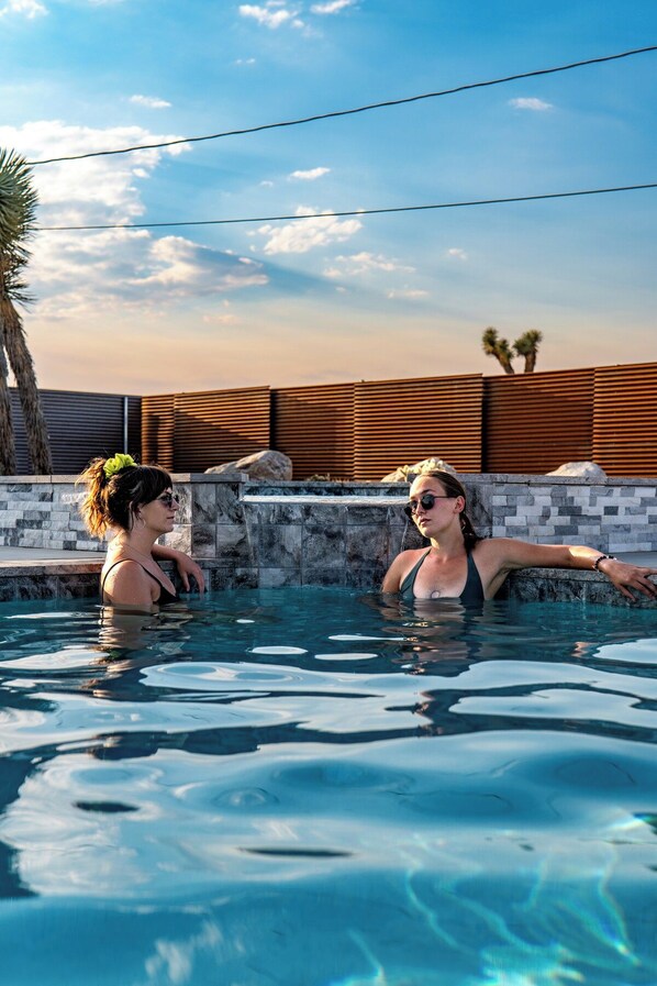 A swimming pool with two people in it, surrounded by a stone wall and a wooden fence.