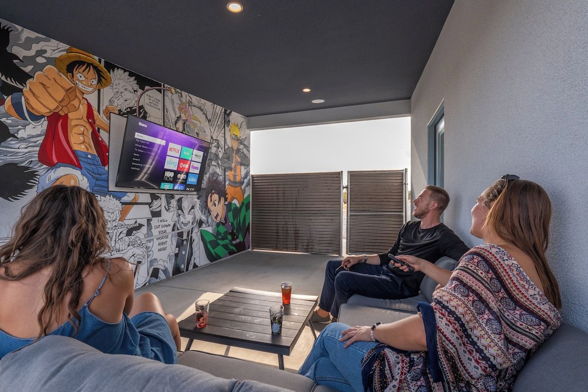 Un groupe de personnes est assis sur un balcon. On peut voir un écran de télévision qui affiche l’interface d’un service de diffusion en continu, une fresque murale représentant des personnages d’anime et une vue sur le ciel.