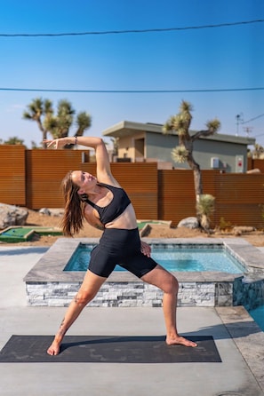 Eine Person macht eine Yoga-Pose draußen, im Hintergrund ein Pool und ein modernes Haus.