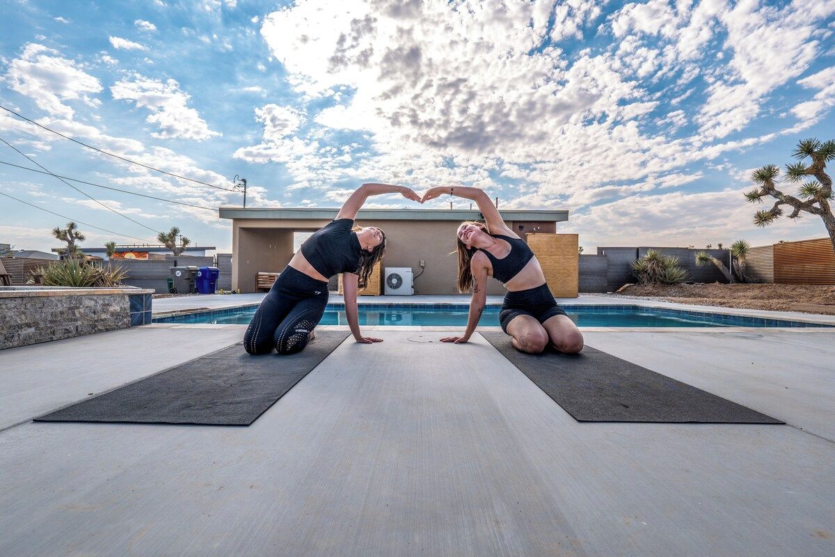 Deux personnes pratiquent une posture de yoga sur des tapis, près d’une piscine, avec un paysage désertique en arrière-plan.