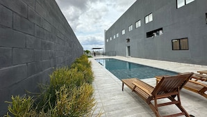 Un espace piscine extérieur moderne, doté d’un mur en béton, d’un espace salon couvert et offrant une vue dégagée sur le ciel.