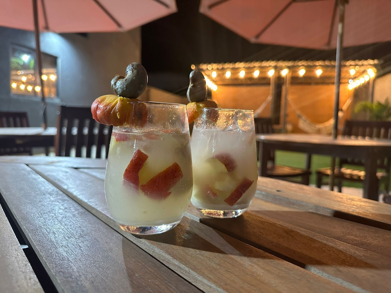 Two glasses of a clear beverage with fruit pieces and a nut on the rim, placed on a wooden table outdoors with string lights in the background.