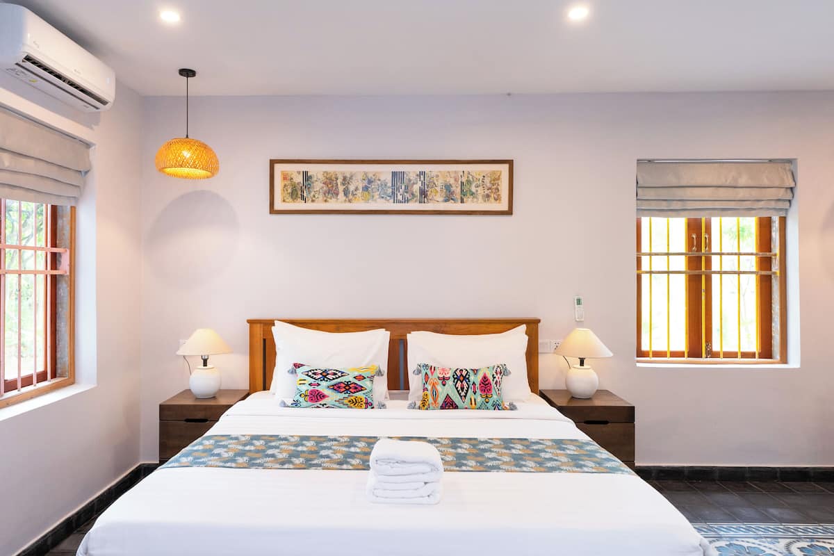 Una habitación de hotel con una cama grande, dos mesitas de noche con lámparas, un ventilador de techo y una ventana con barrotes.