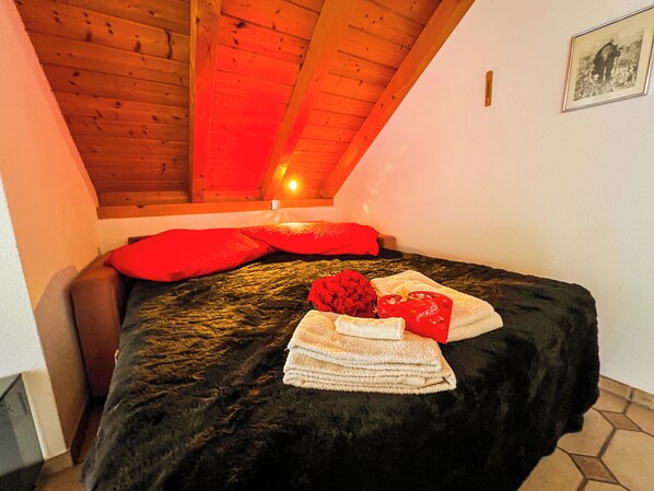 Un dormitorio con techo inclinado, cama con colcha negra, almohadas rojas y una pila de toallas.