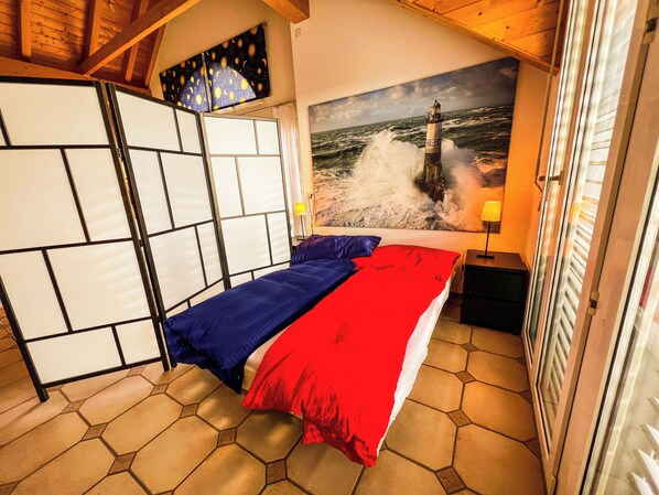 Un dormitorio con cama, mesita de noche, lámpara, un cuadro de un faro y vista al mar.
