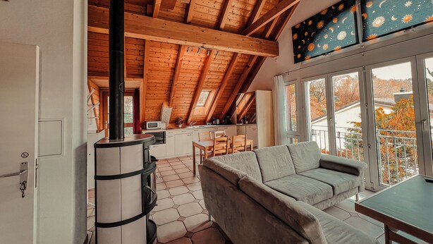 Ein gemütliches Dachzimmer mit Holzbalken, einer Couch, einem Esstisch und einem großen Fenster mit Blick auf Bäume.
