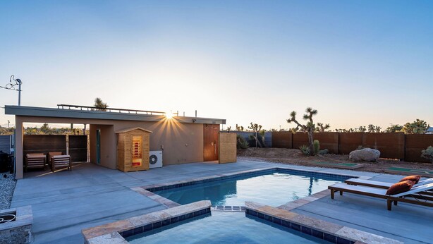 A modern outdoor pool area with a sunlit patio, lounge chairs, and a small building with a door. - Tiny Home - Salt Water Pool - Jacuzzi - Mini Golf - Sauna (Yucca Valley)