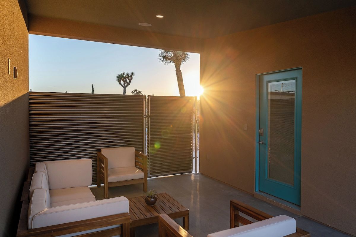 Un espace extérieur aménagé avec un canapé et des fauteuils, une table agrémentée d’une plante, et une porte donnant sur une vue de palmiers et du coucher de soleil.