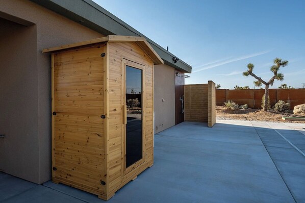 A wooden structure with a sliding door, adjacent to a building with a sloped roof and a concrete patio. - Tiny Home w Pool, Hot Tub, Sauna, Golf & More! (Yucca Valley)