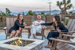 Three people seated around a fire pit in an outdoor setting with a view of mountains.
