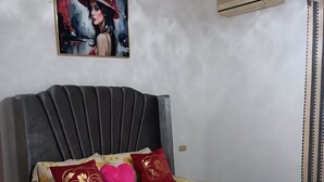 Un dormitorio con cama, cabecera, aire acondicionado de pared, un cuadro y una cortina.