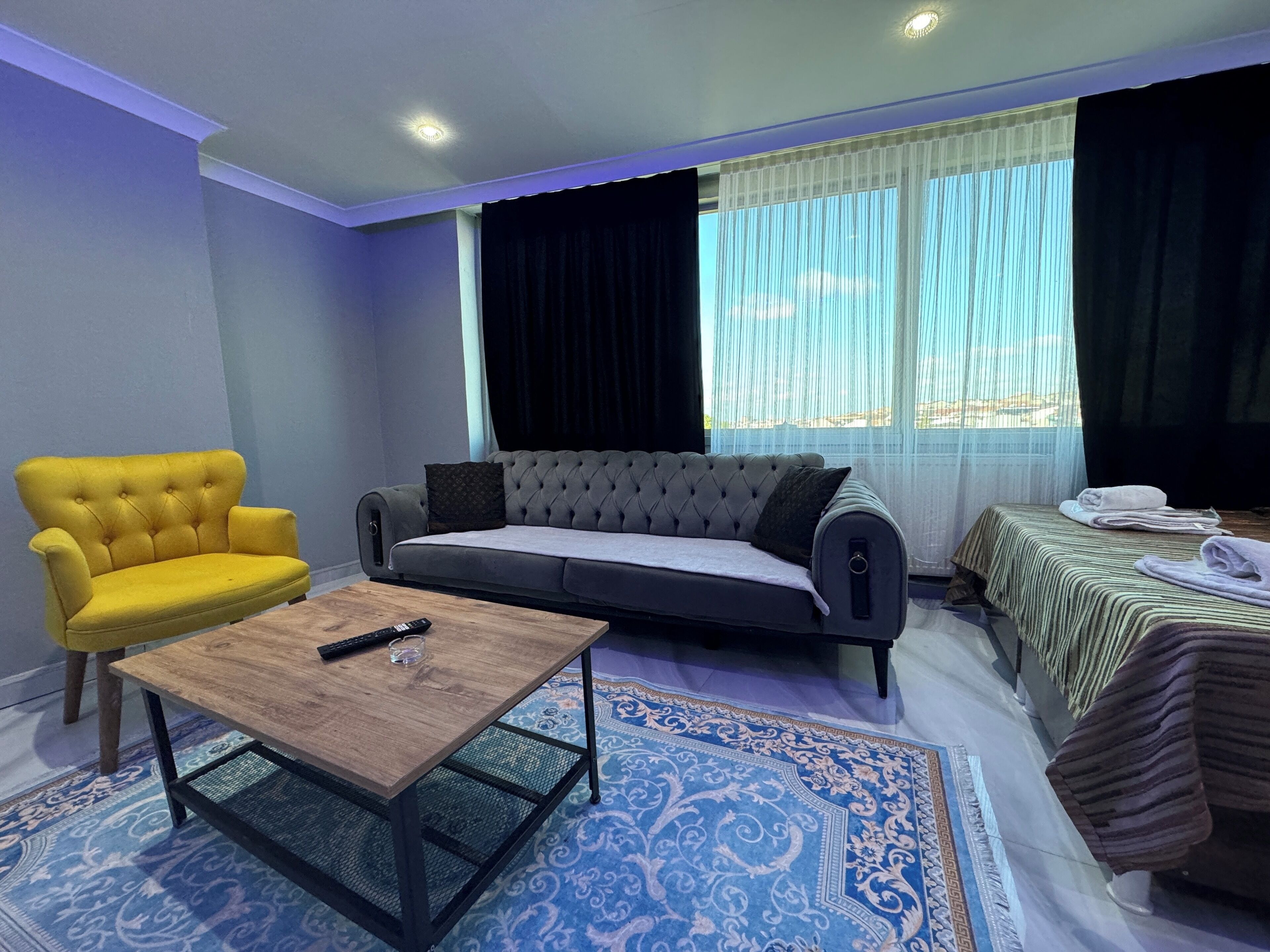 A living room with a yellow armchair, a grey sofa, a wooden coffee table, and a bed with a striped bedspread.