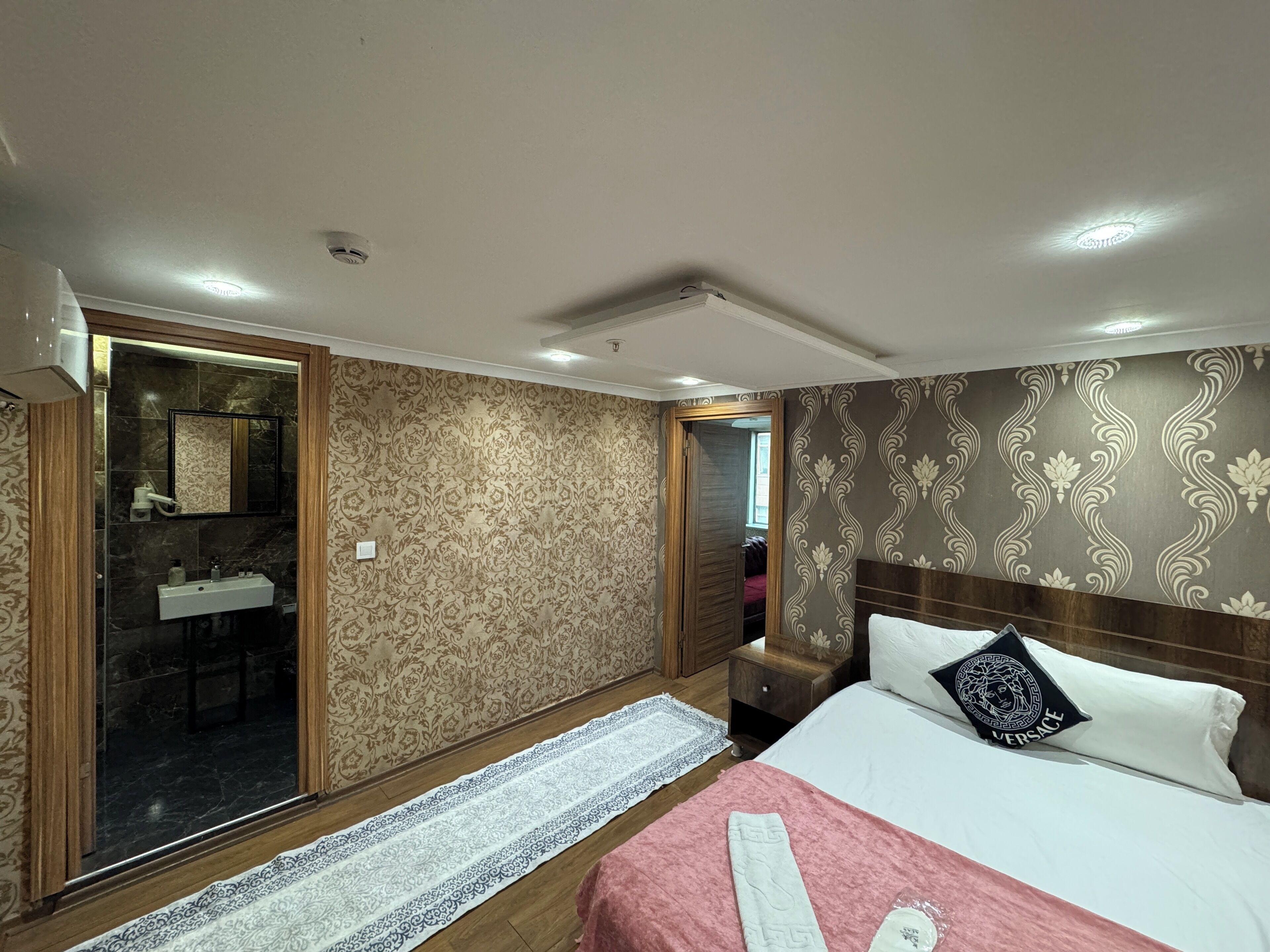 A bedroom with a bed, a bedside table, a nightstand, a mirror, a bathroom with a sink, and a patterned wall.
