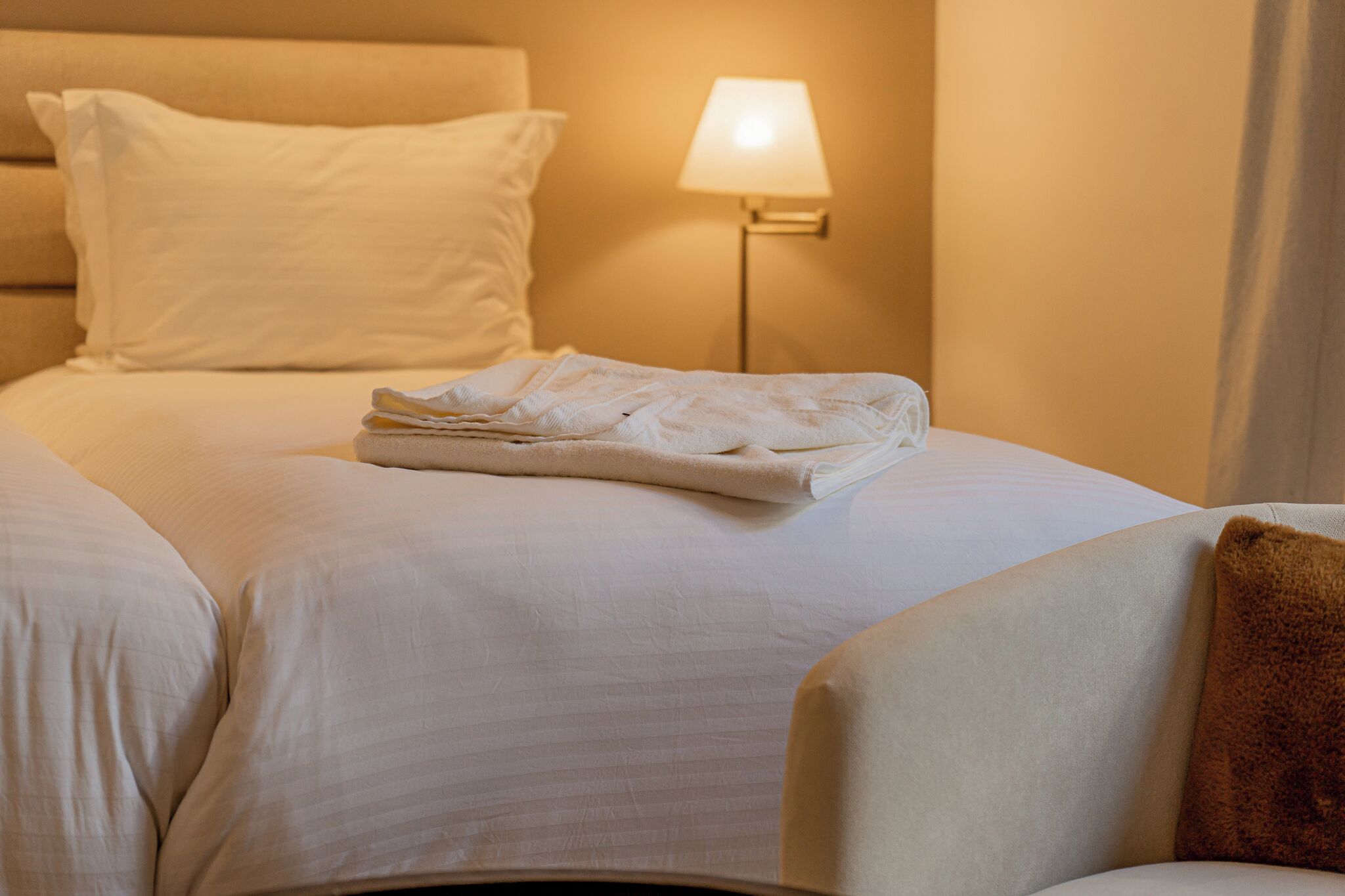 A neatly made bed with white linens and a folded towel on top, a bedside lamp, and a brown pillow on the right side.