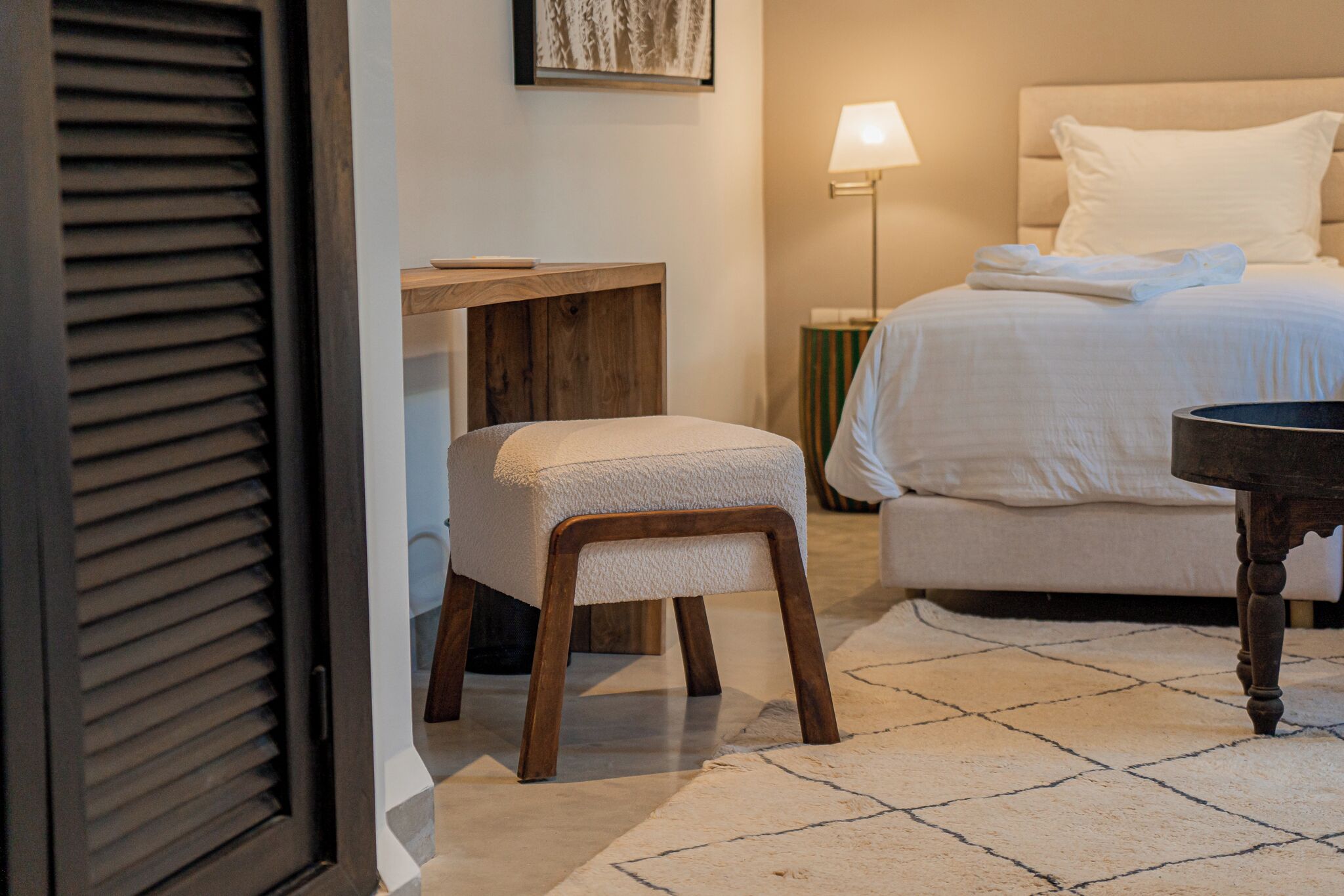 A hotel room with a bed, a bedside table, a chair, a lamp, and a door with blinds.