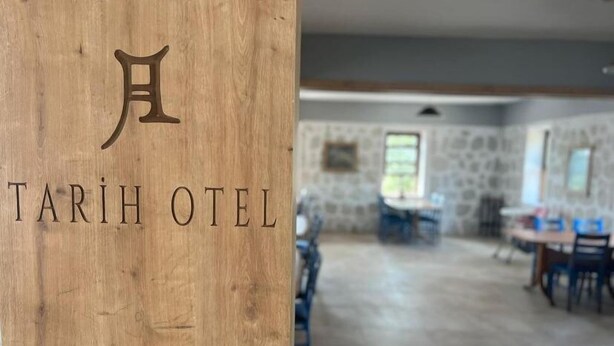 Un letrero de madera con el nombre "TARİH OTEL" y vista de una habitación amplia con paredes de piedra y un área de comedor.