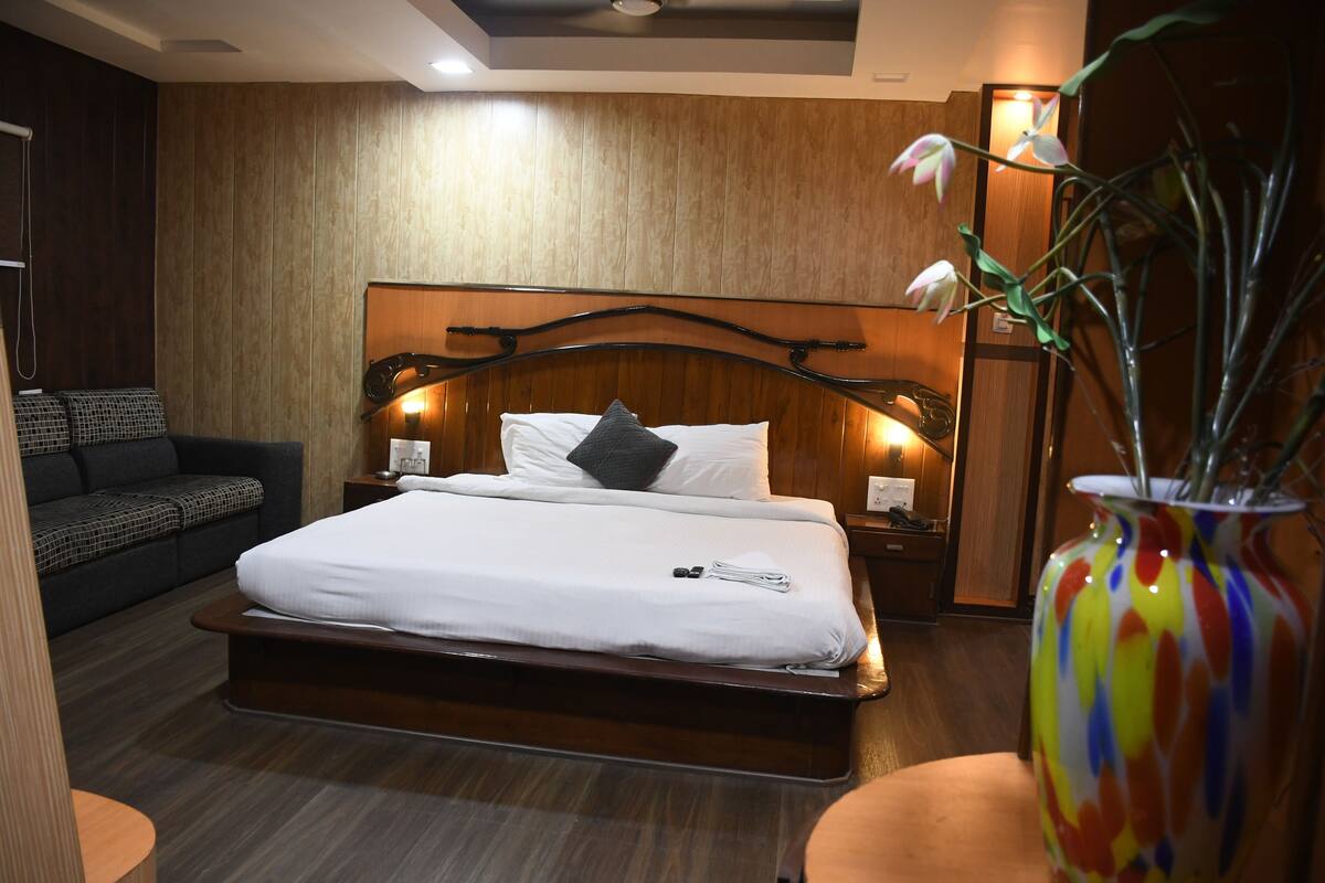 Ein modernes Hotelzimmer mit einem großen Bett, einem Holz-Kopfteil, Nachttischen mit Lampen, einem dunkelgrauen Sofa und einer farbenfrohen Vase auf einem Beistelltisch.