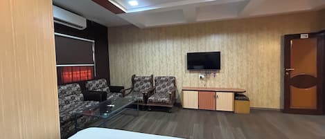 A hotel room with a bed, a glass table, two patterned armchairs, a TV mounted on the wall, and a wooden cabinet.