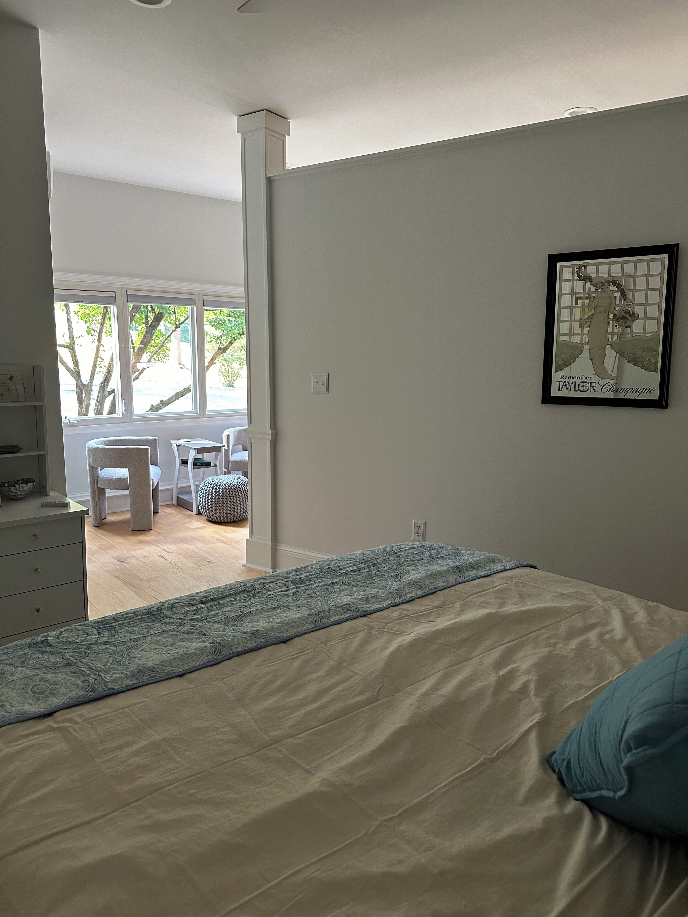Une chambre avec un grand lit, un bureau et une chaise. Il y a une fenêtre donnant sur des arbres et un balcon.