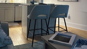 Une cuisine moderne avec des meubles blancs, un îlot central doté de tabourets de bar et un éclairage suspendu. On y trouve un coin repas avec un canapé et une table, ainsi qu’un grand tableau abstrait accroché au mur.