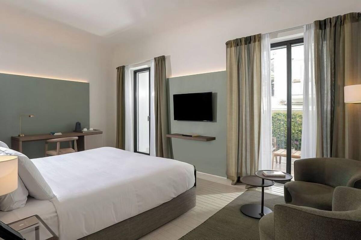 A hotel room with a large bed, a desk with a chair, a TV mounted on the wall, and a view of the outdoors through the window.