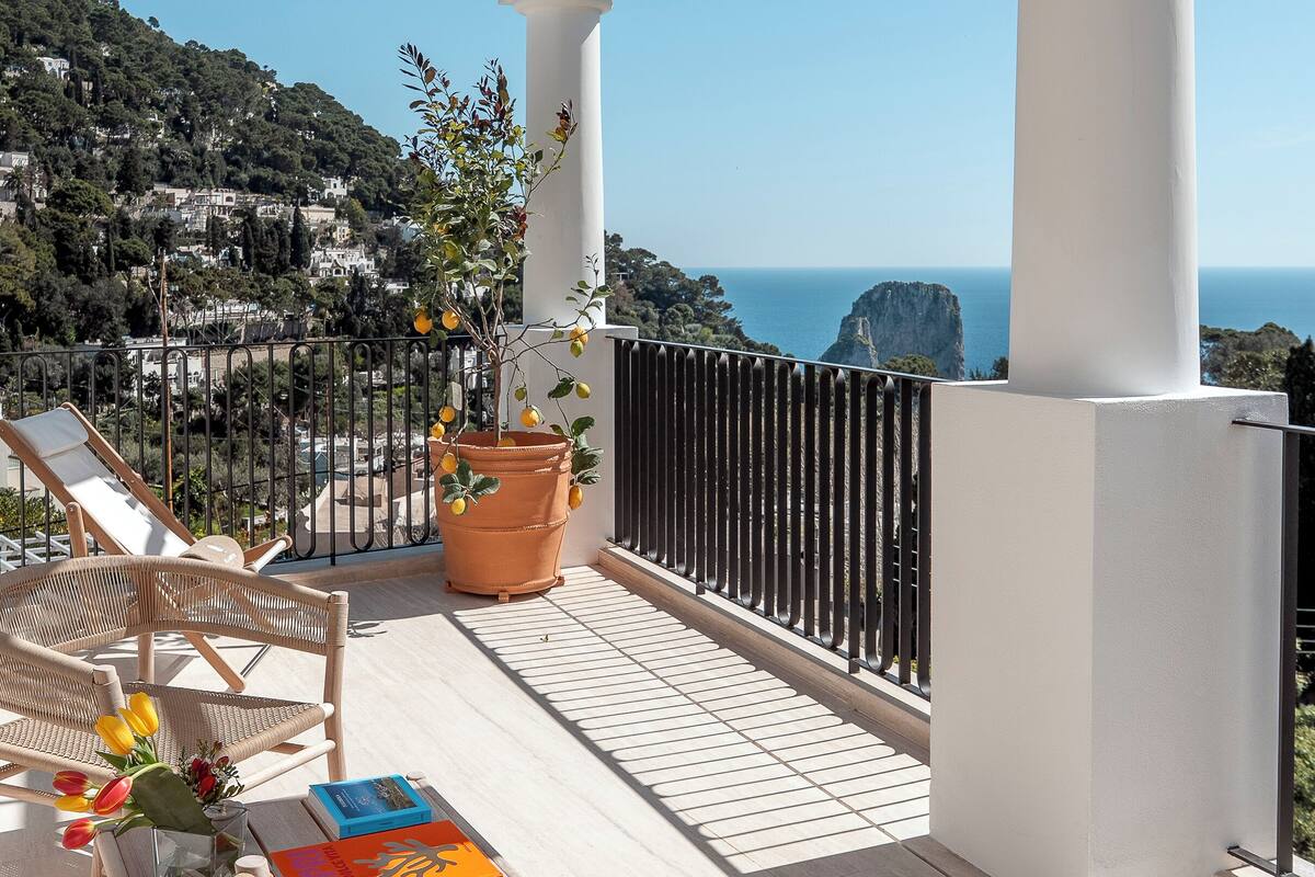 A balcony with wicker chairs, a table with books, a potted plant, and a view of the sea and hills.