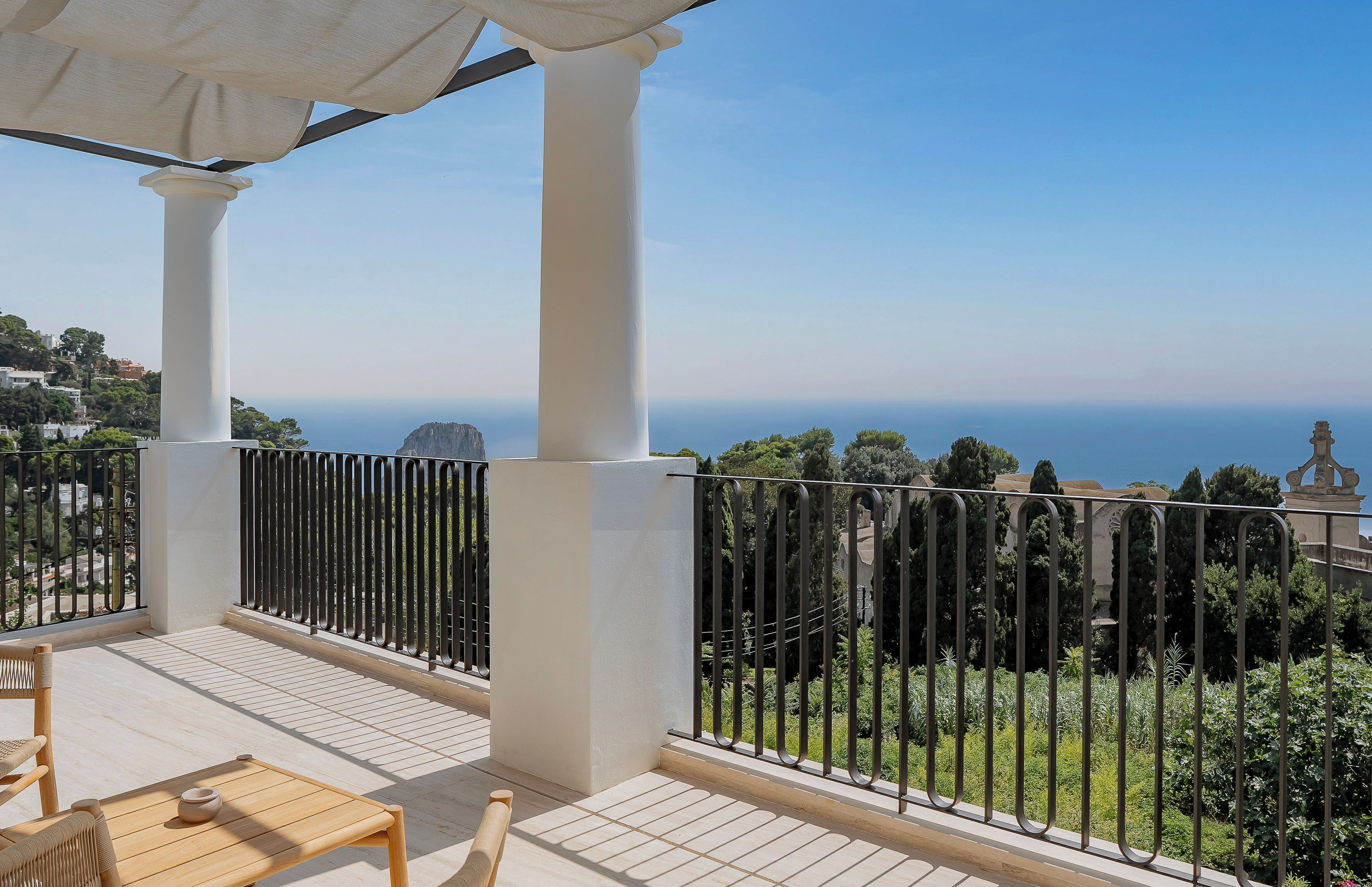 A balcony with a view of the sea, white columns, and a table with a teapot.