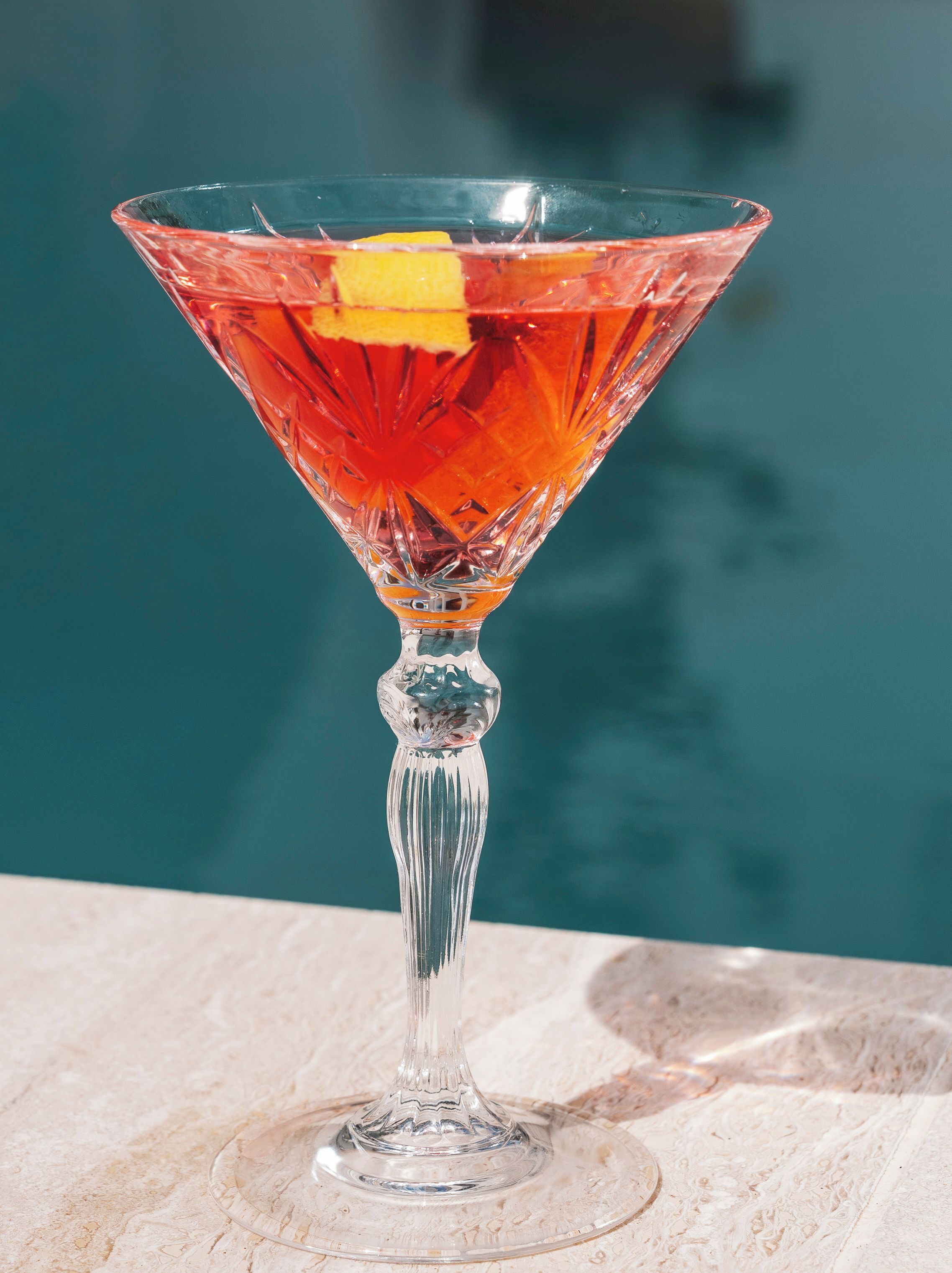 A clear glass cocktail with a lemon slice garnish, placed on a marble surface near a pool.