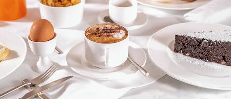 A breakfast setting with a coffee cup, a chocolate cake slice, and various breakfast items on a table.