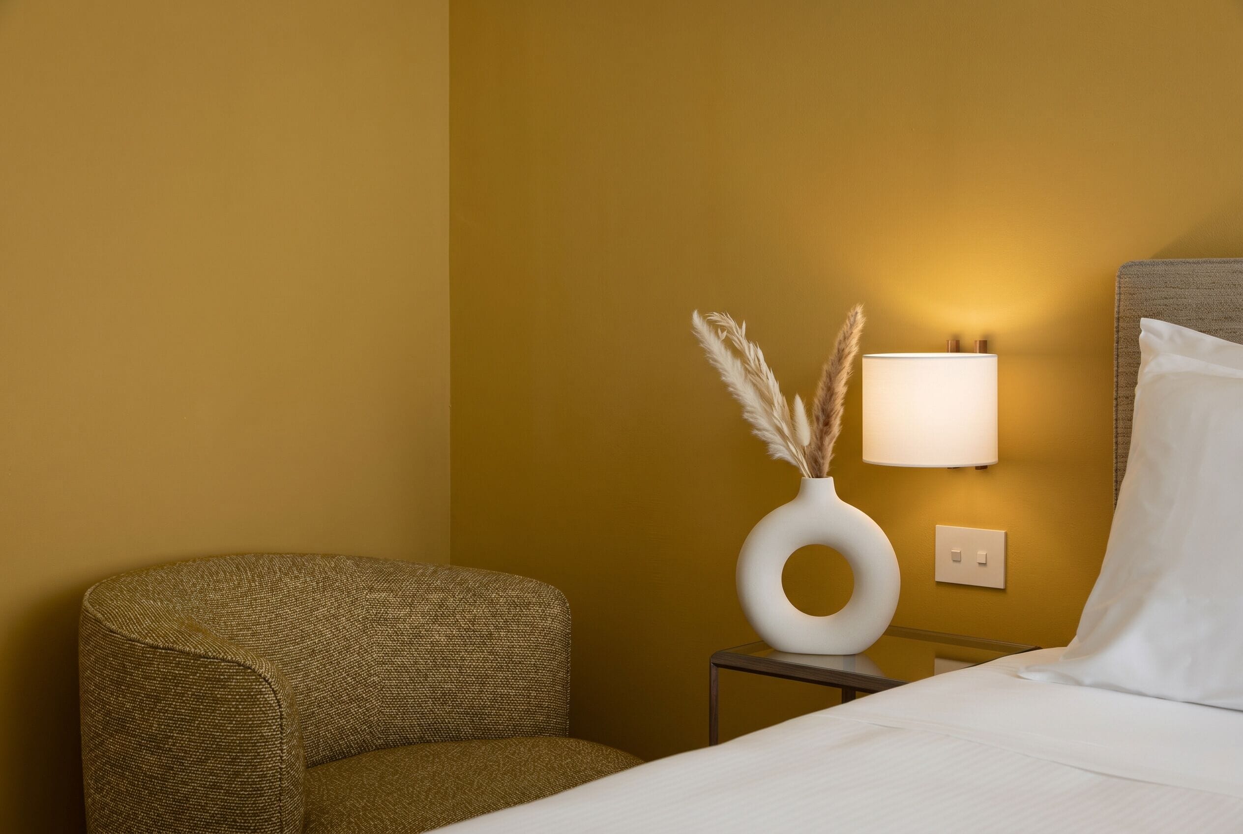 Una habitación de hotel con una cama, una mesita de noche con lámpara, un jarrón con plumas de pampa y un sillón con textura.