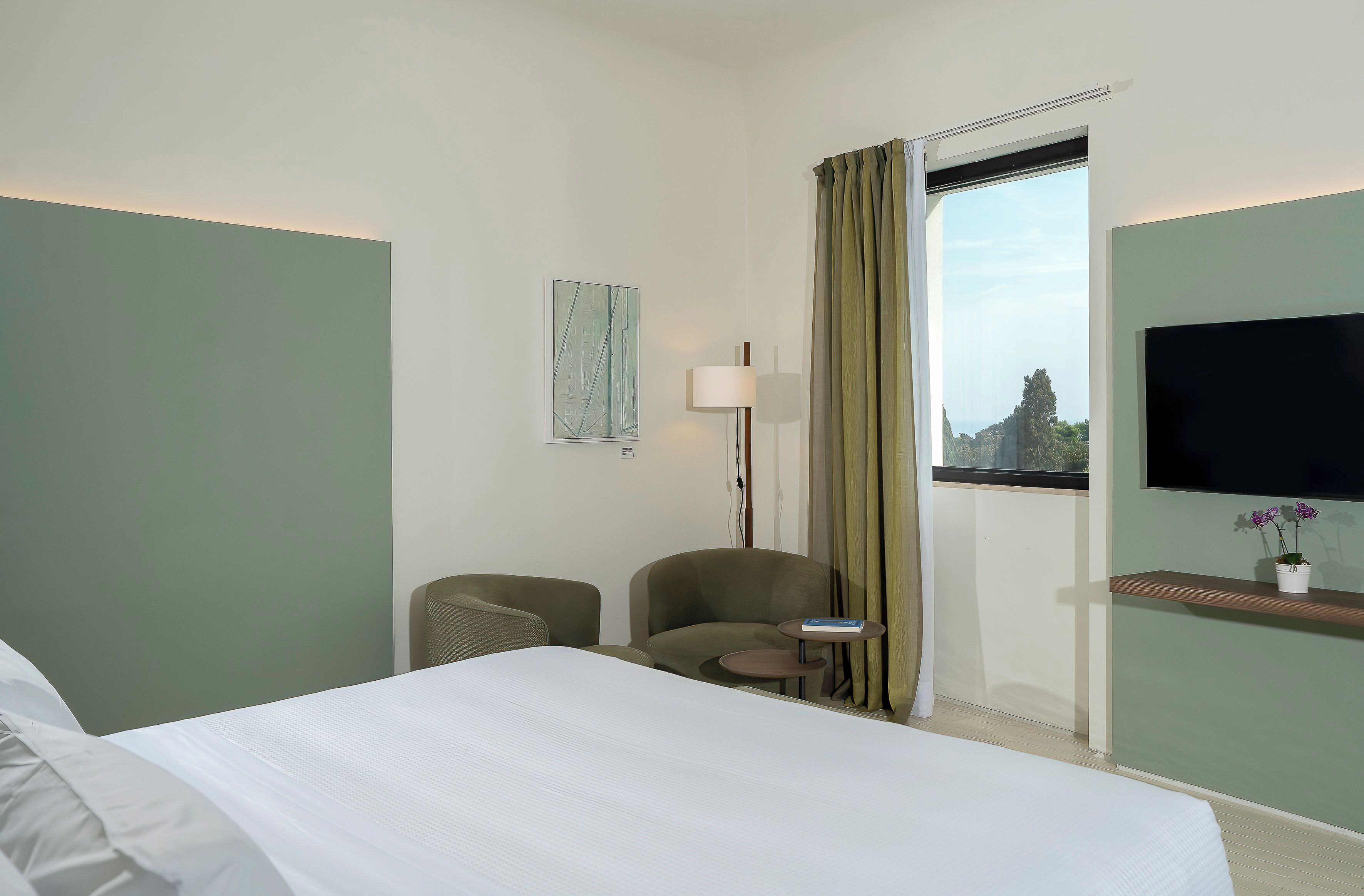 A hotel room with a bed, two chairs, a small table, a lamp, a TV, and a window with curtains.