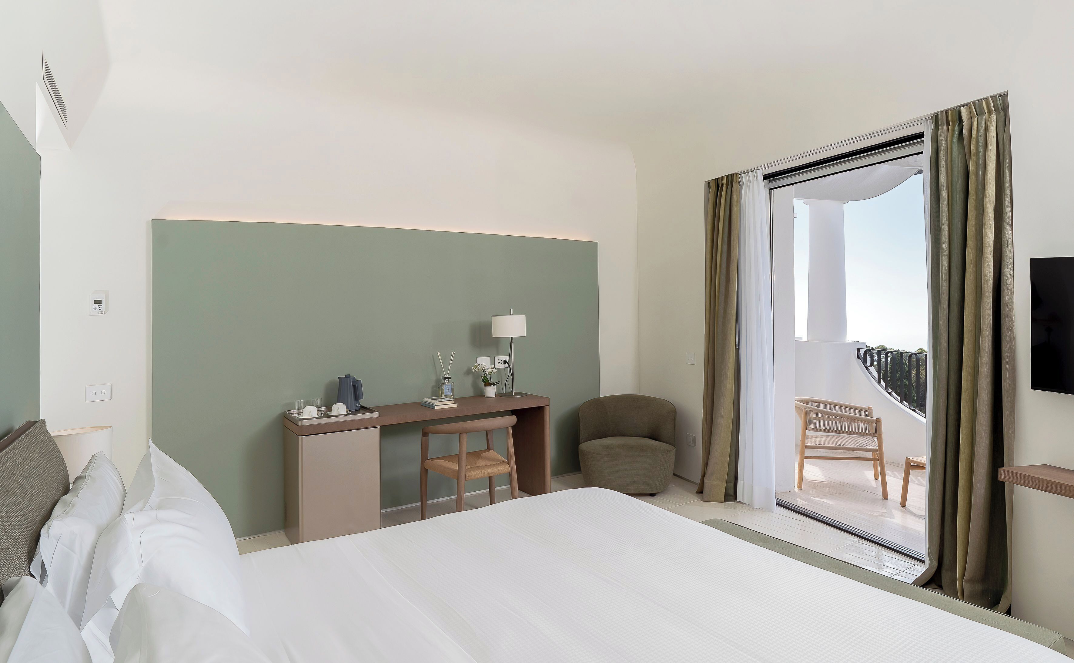 Ein Hotelzimmer mit Bett, Schreibtisch mit Lampe, Stuhl und einem Balkon mit Blick.