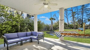 A covered patio with a blue sofa, a hammock, and a view of a lush garden and trees. - 4BR/3BA Retreat - Perfect for Families & Groups (Tybee Island)