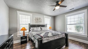 Un dormitorio con cama, dos mesitas de noche, cómoda y ventilador de techo, junto con ventanas amplias con persianas.