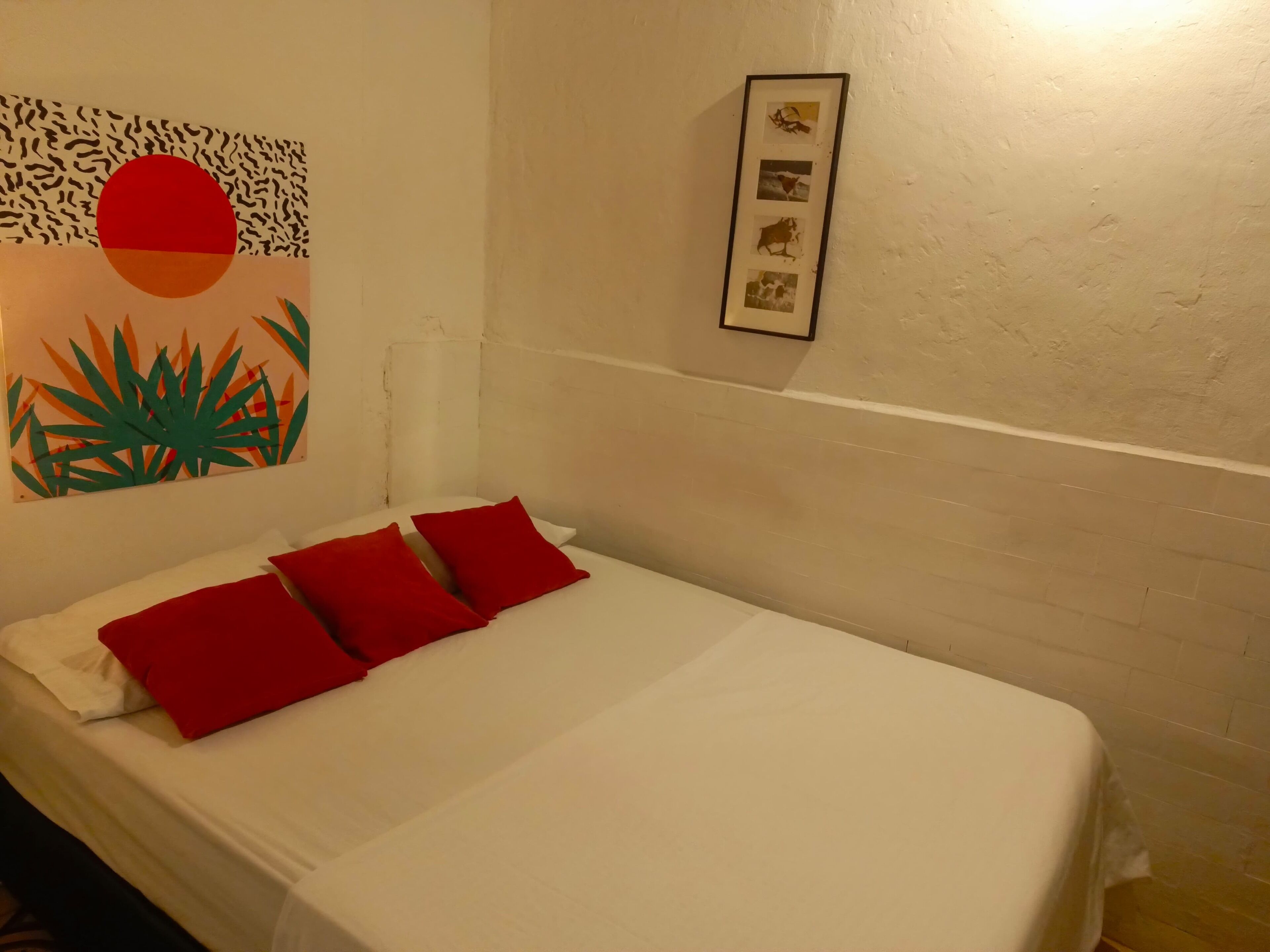 Un lit bien fait, agrémenté d’oreillers rouges, une œuvre d’art murale représentant un soleil et des plantes, et une collection de photos encadrées accrochées au mur.