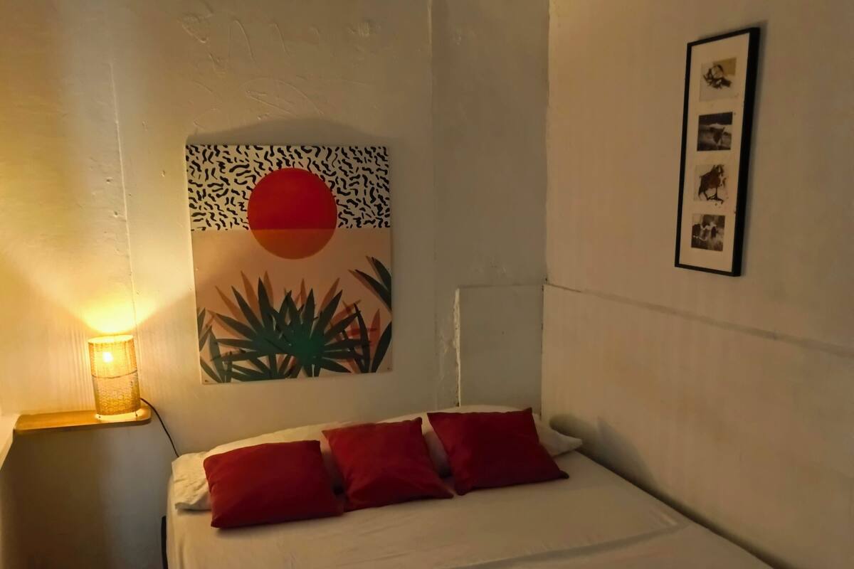 Una cama bien tendida con ropa de cama blanca y roja, un cuadro en la pared con un sol y follaje, y una fotografía enmarcada sobre la cama.
