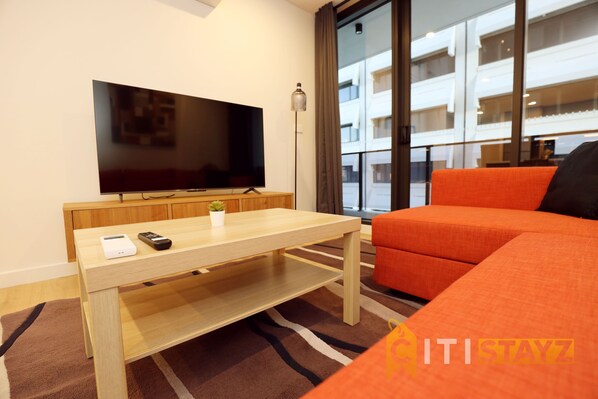 A modern living room with a flat-screen TV, a wooden coffee table, a small plant, and an orange sofa.