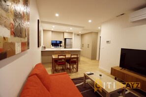 A modern living room with a sofa, a coffee table, and a TV. In the background, there is a kitchen with a dining area.