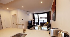 A modern kitchen with a sink, toaster, and kettle, a dining area with a glass table, and a view of the city through large windows. - Red Rose Retreat - 1bd 1bth Apt (Phillip)